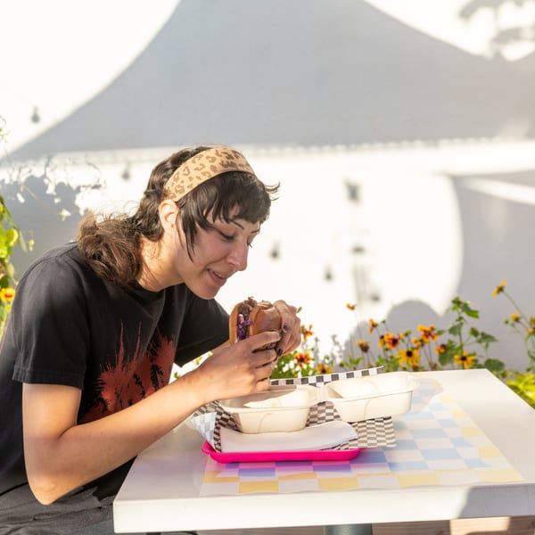 A person in a black shirt and leopard print headband eats a burger at an outdoor table.