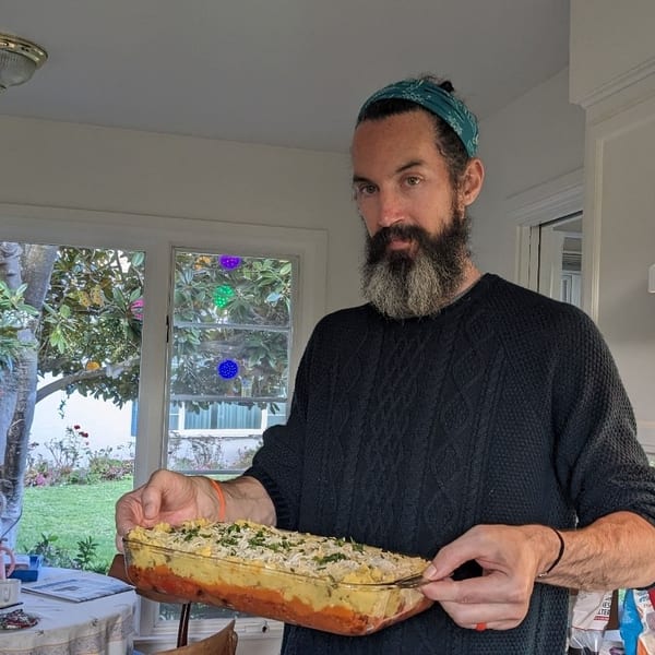 A man in a kitchen holds a glass baking dish filled with a layered casserole.