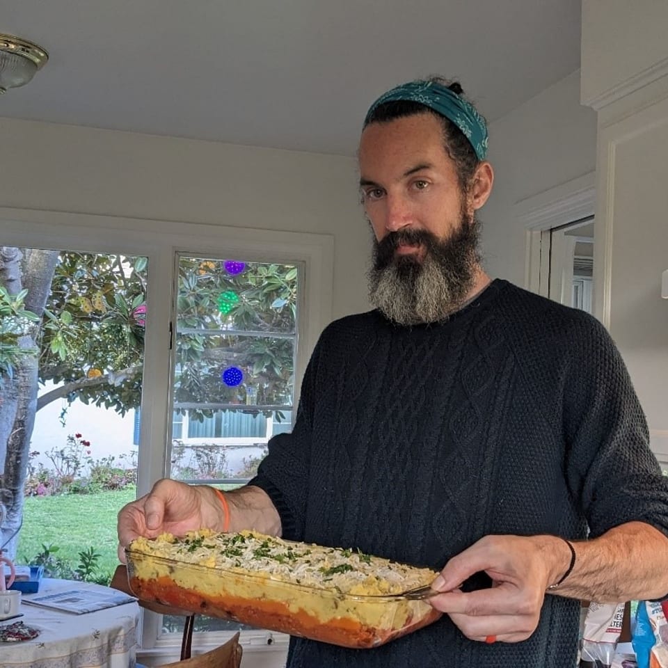 A man in a kitchen holds a glass baking dish filled with a layered casserole.