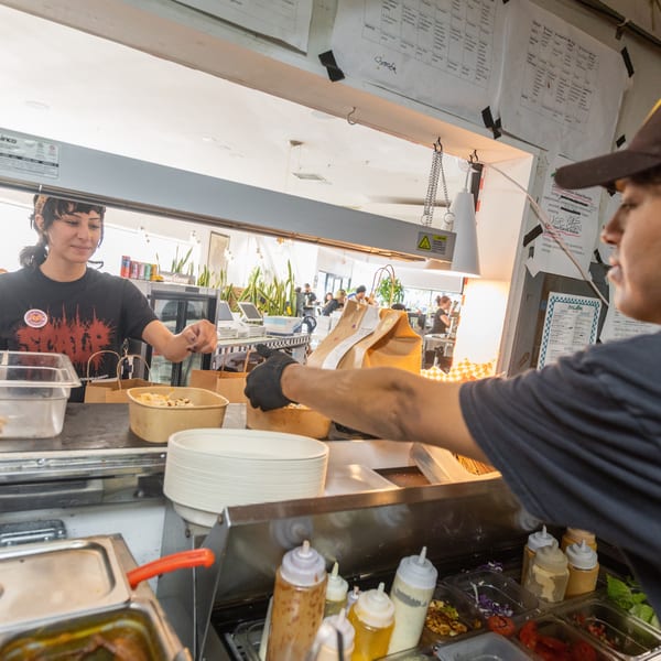 Two restaurant workers preparing food and packaging orders at a service counter with sauces and ingredients.