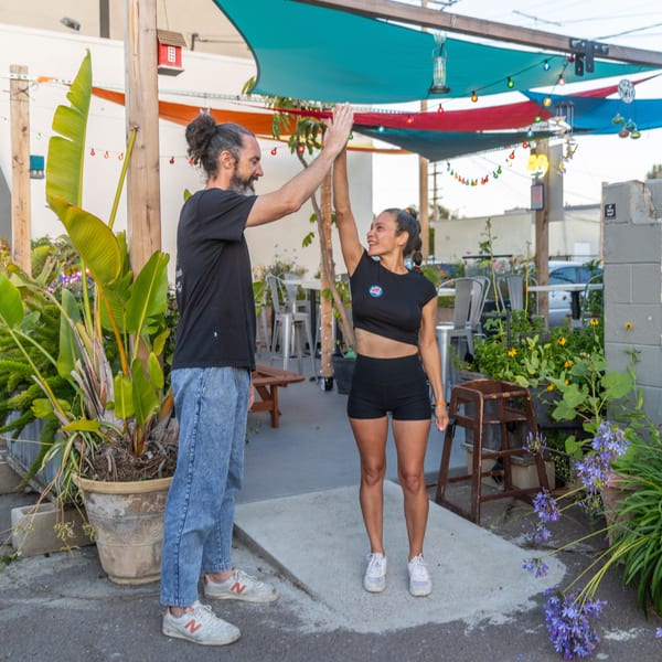 Two people high-fiving outdoors under colorful shade sails, surrounded by lush plants and string lights.