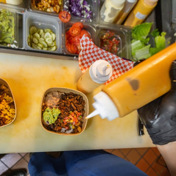 Chef in black gloves squeezing sauce onto a bowl of food with various toppings in containers.