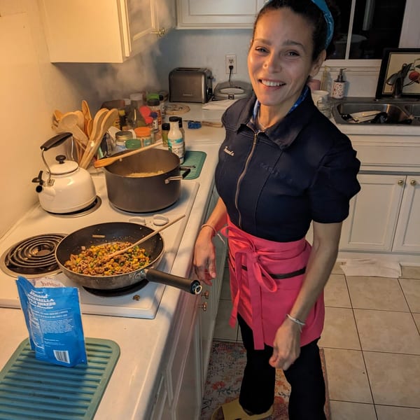 A smiling woman in a blue top and pink apron cooks in a kitchen with steaming pots on the stove.