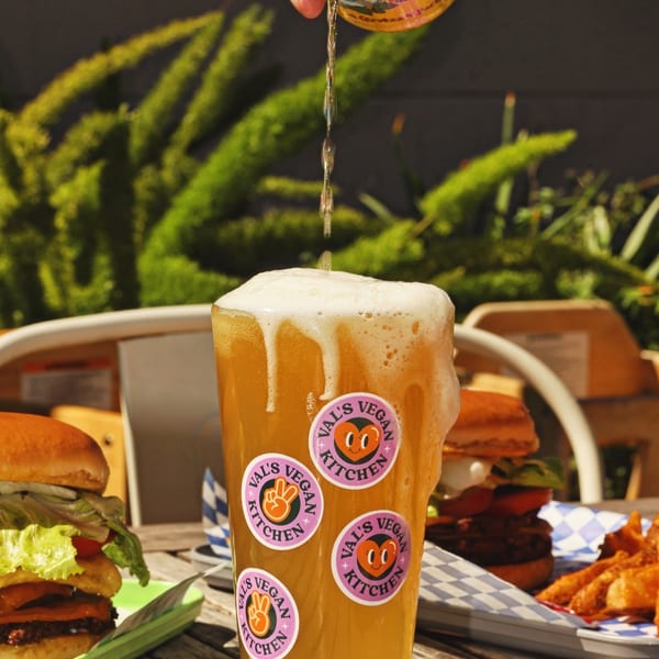 Beer being poured into a Val's Vegan Kitchen glass, with burgers and fries on a wooden table.