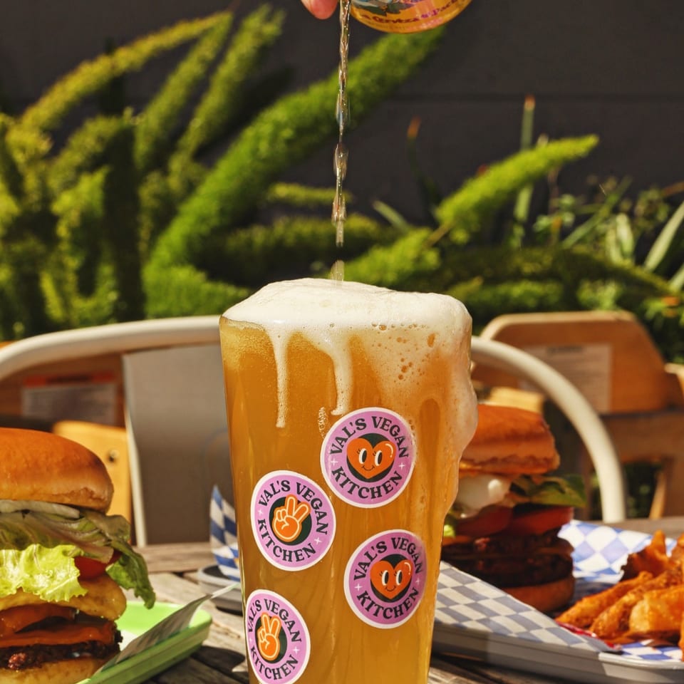 Beer being poured into a Val's Vegan Kitchen glass, with burgers and fries on a wooden table.