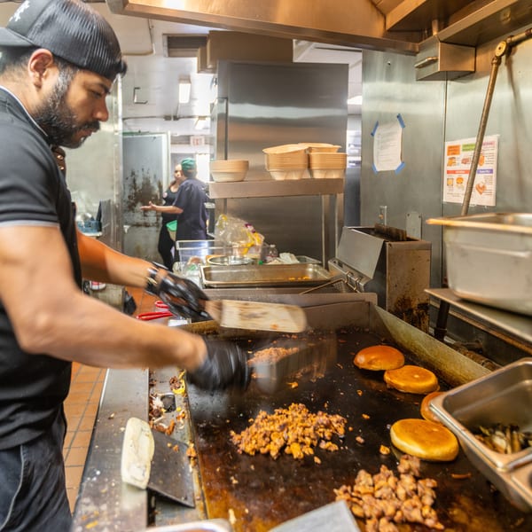 Chef in a black cap and shirt cooking meat and buns on a griddle in a busy kitchen.