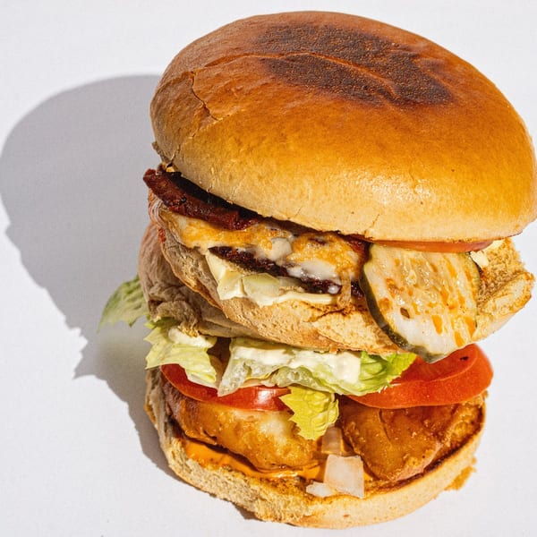 Tall double-decker burger with cheese, bacon, pickles, lettuce, tomato, and sauce on a white background.