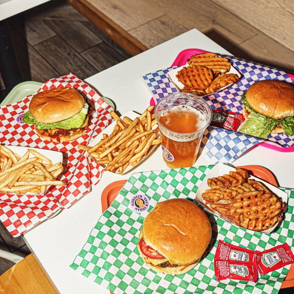 Three burgers, two with fries, one with waffle fries, and a beer on a white table.