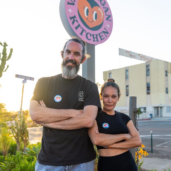 Two smiling people, a man and a woman, stand with crossed arms in front of a "Vegan Kitchen" sign.
