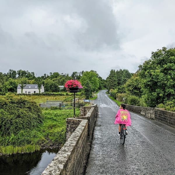 Person in a pink Nirvana jacket cycling over a stone bridge in a lush green rural landscape.